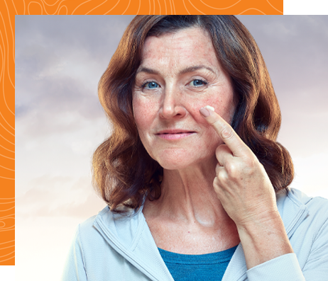 A middle-aged woman with brown hair and rosacea looks at the camera while applying foam to her face with one finger